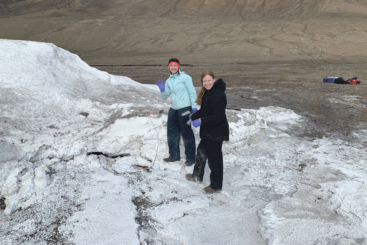 Photo de deux femmes habillées chaudement qui se tiennent debout sur un sol blanc. À l'arrière-plan, des pentes de sol brun dénué de végétation.