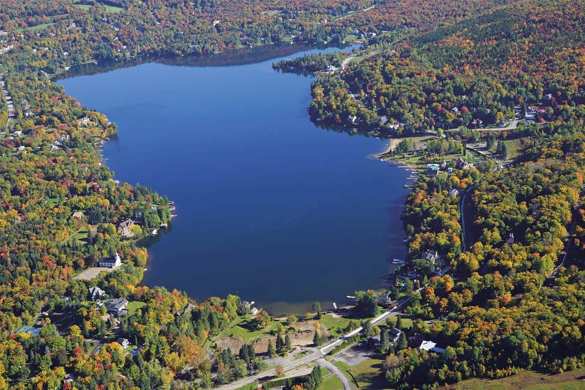 Photo aérienne du Lac Beauport prise au début de l'automne. Tout bleu au centre de l'image. Autour, des secteurs boisés avec des feuillus qui commencent à avoir des couleurs, une petite route et quelques résidences.