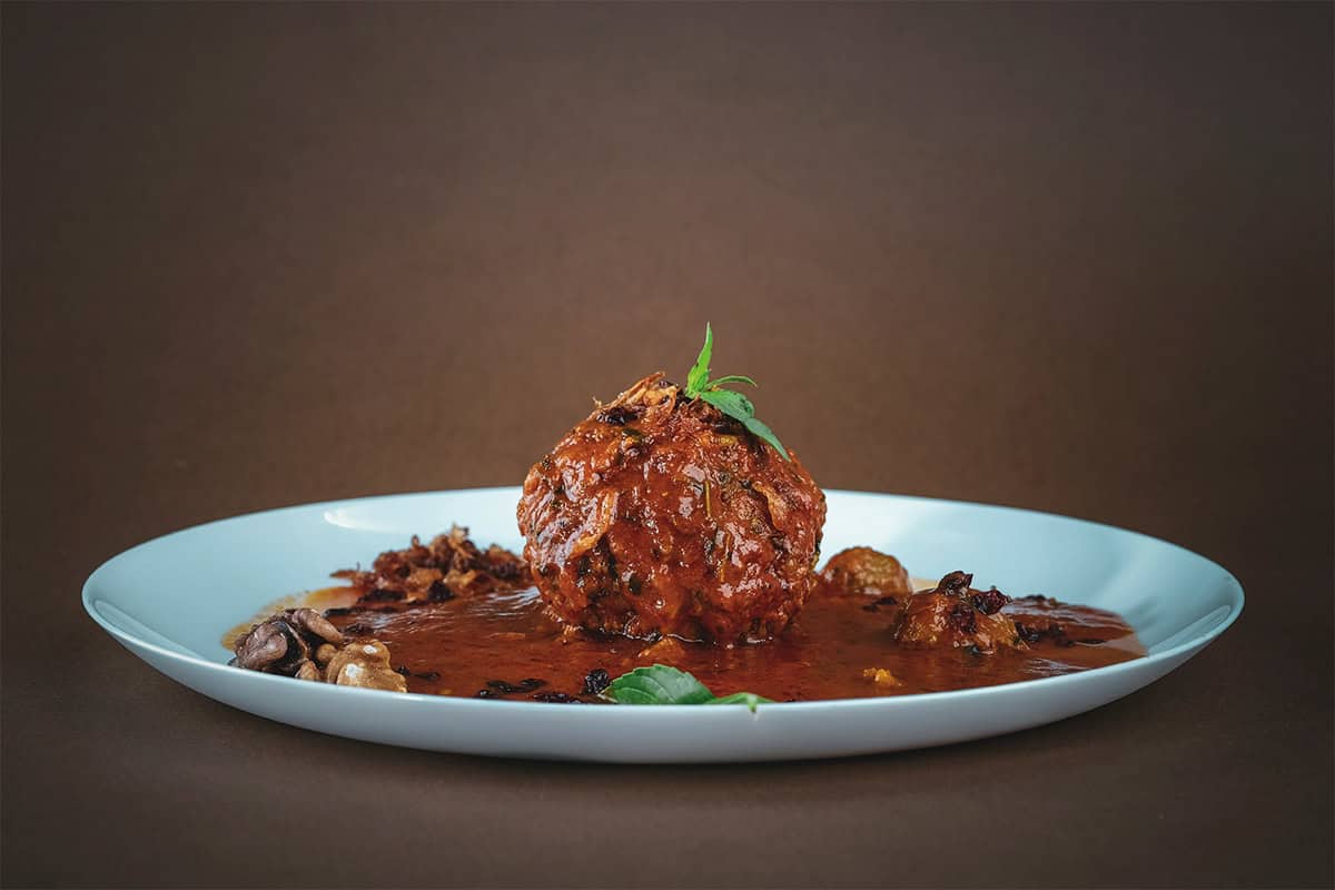 Une boulette de viande cuite déposée au centre d'une assiette en porcelaine blanche. La viande est nappée de sauce et décorée d'herbes fraîches.