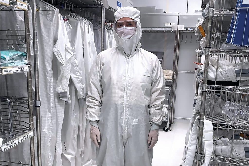 Une femme portant une combinaison blanche à capuchon, un couvre-visage, et des lunettes de sécurité transparentes. Elle se tient debout dans un un vestiaire. Derrière elle, d'autres combinaisons blanches sont accrochées sur une tringle métallique..