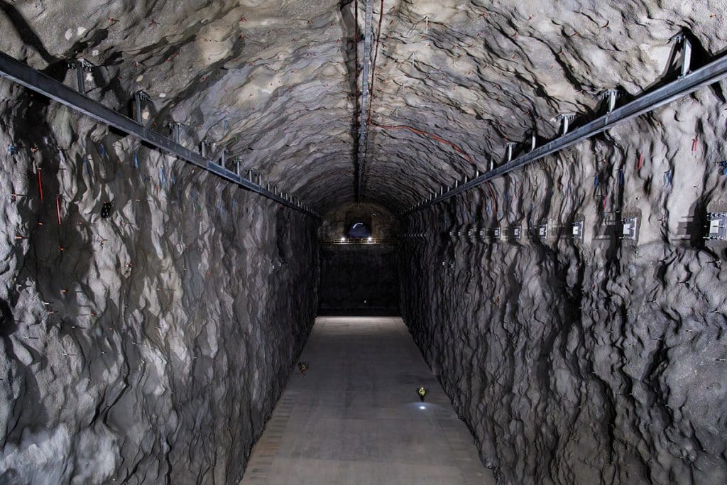 Photo: Un tunnel très droit creusé dans la roche. Des rails de métal sont fixés au plafond.