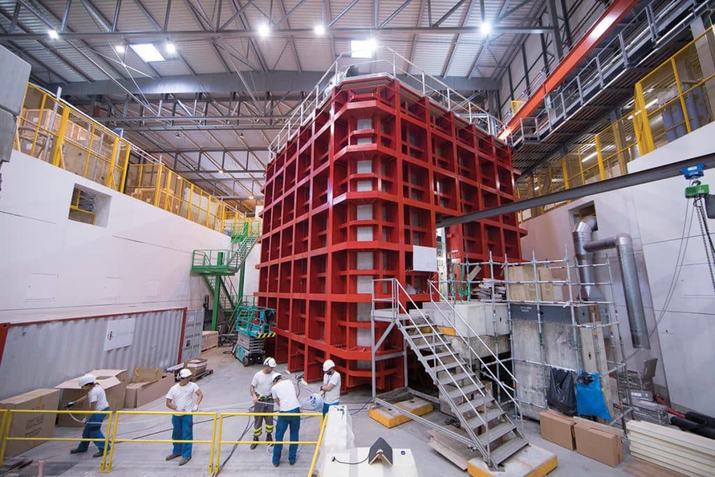 Une grande salle haute de plusieurs étages. 5 personnes s'affairent autour d'une grande installation.