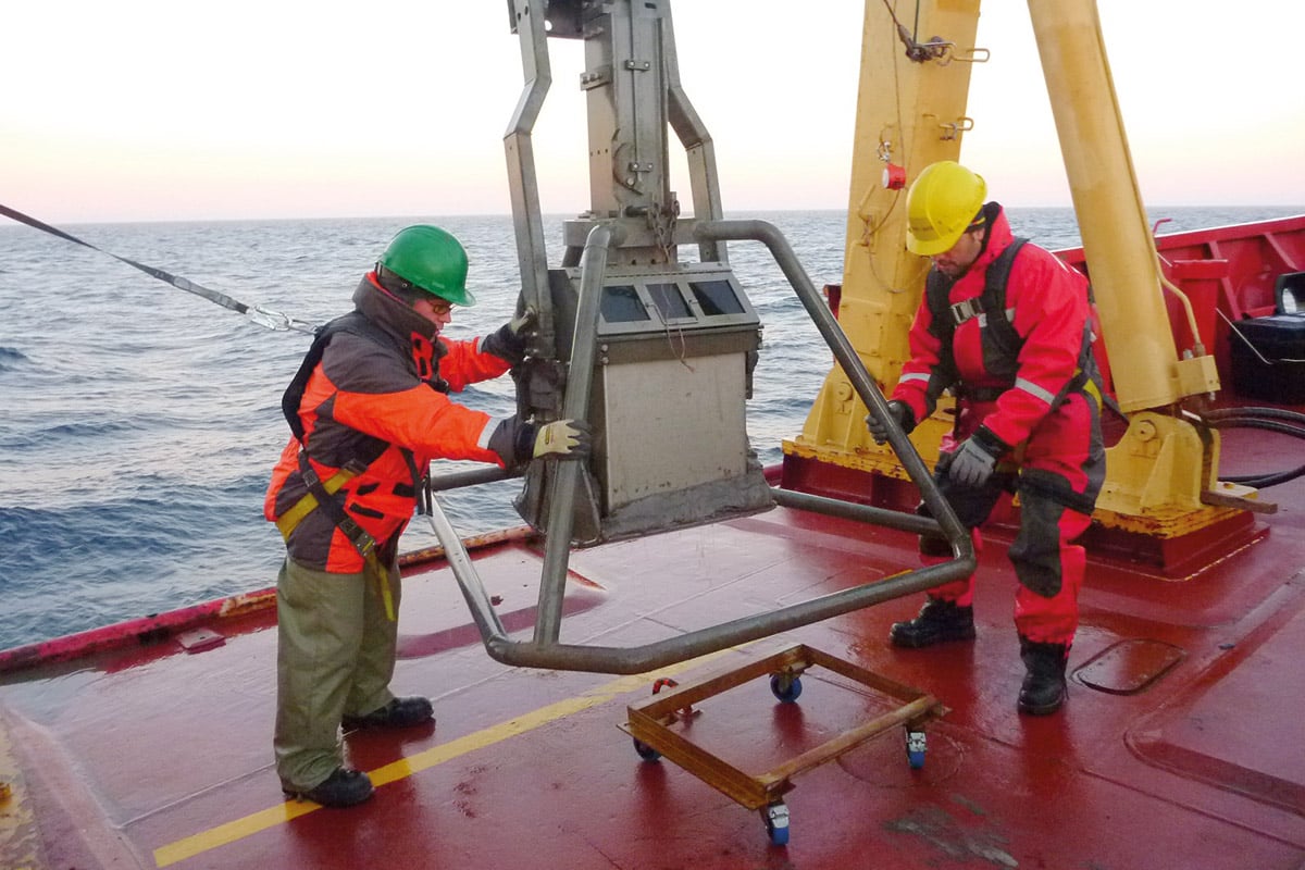 Photo: Deux hommes portant des manteaux chauds, des bottes de travail et un casque de sécurité sont debout sur le pont d'un navire de recherche. Ils manipulent un gros équipement de métal. À l'arrière-plan, on aperçoit la surface de l'eau à perte de vue.