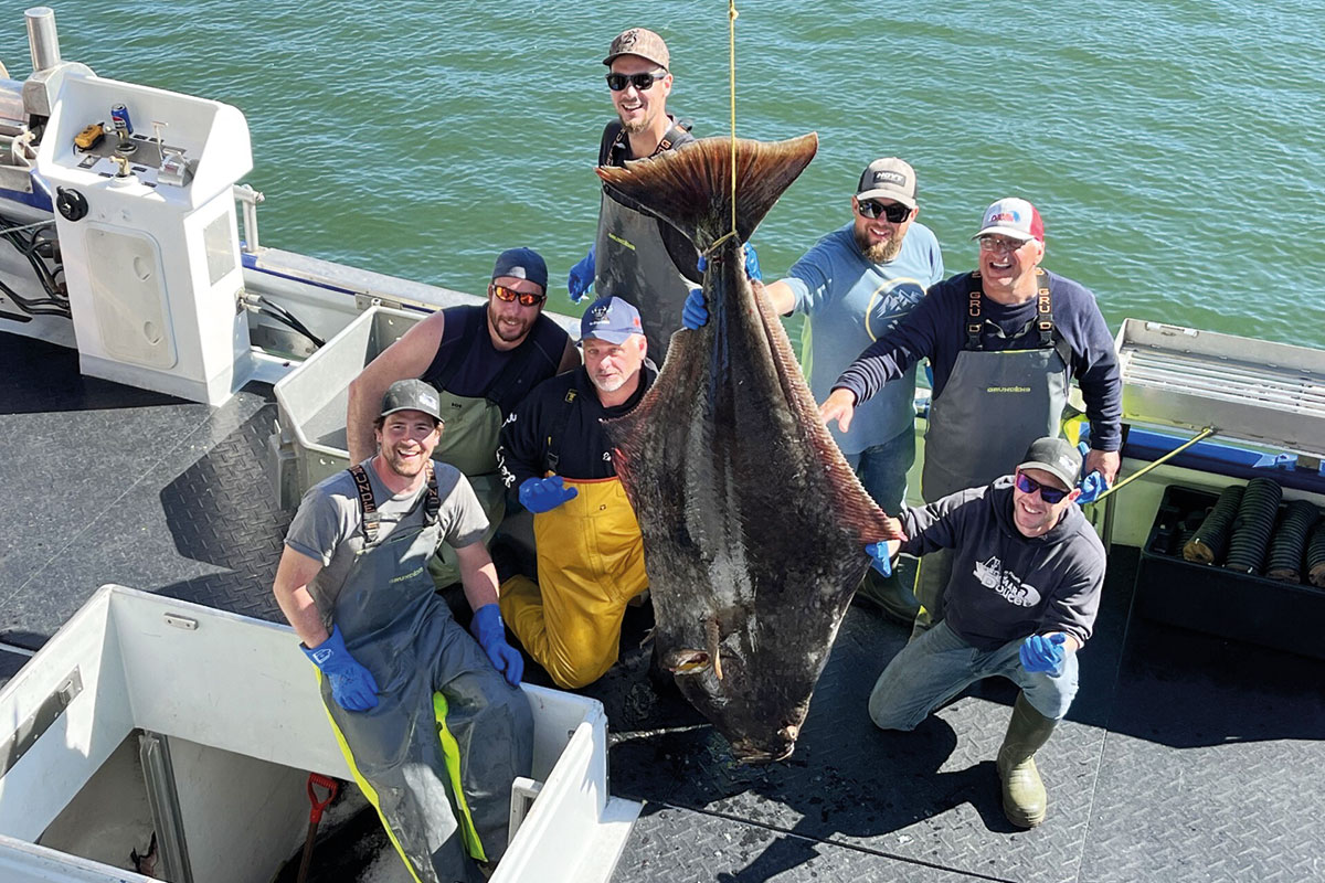 Sur le pont d'un bateau de pêche, 7 hommes en jeans et salopettes de pêche posent fièrement aux côtés d'un très grand poisson suspendu par la queue. Il fait soleil. Quatre hommes portent des lunettes fumées. À l'arrière-plan, la surface de l'eau est calme.