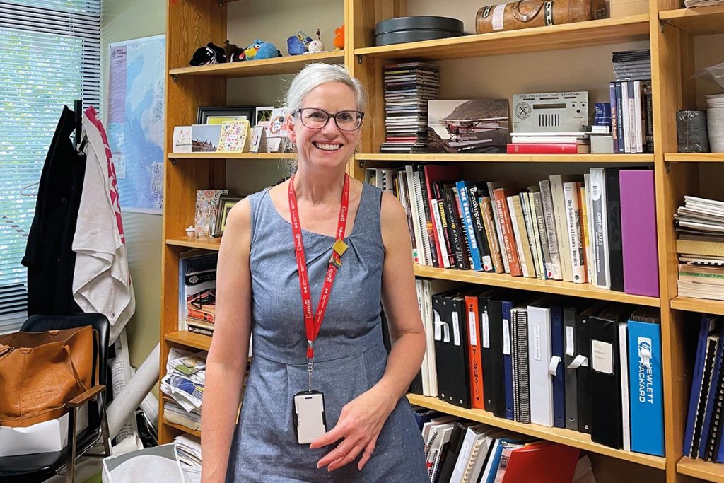 Une femme souriante, portant des lunettes, se tient debout devant des bibliothèques en bois, remplies de livres.
