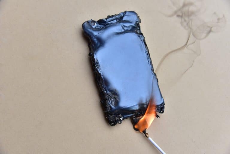 Photo d'un téléphone cellulaire posé sur une surface plante. Il est en train de fondre. Des flammes et de la fumée s'échappent du point de connexion avec le câble de recharge.